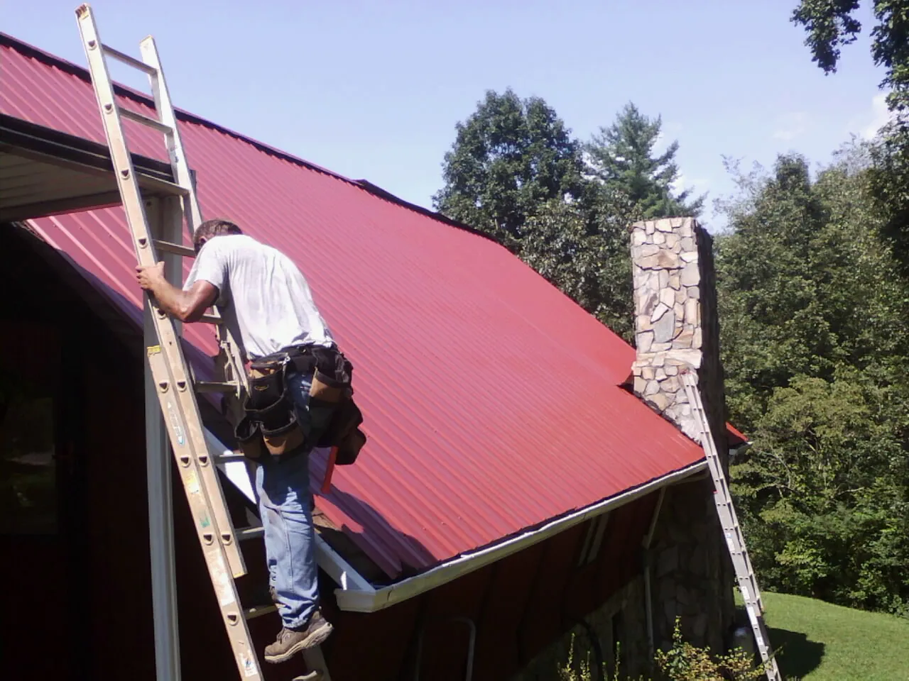 Roofer on a ladder beside a red metal roof and stone chimney — Soffit & Fascia Repair by Roofing Brothers in Scarborough